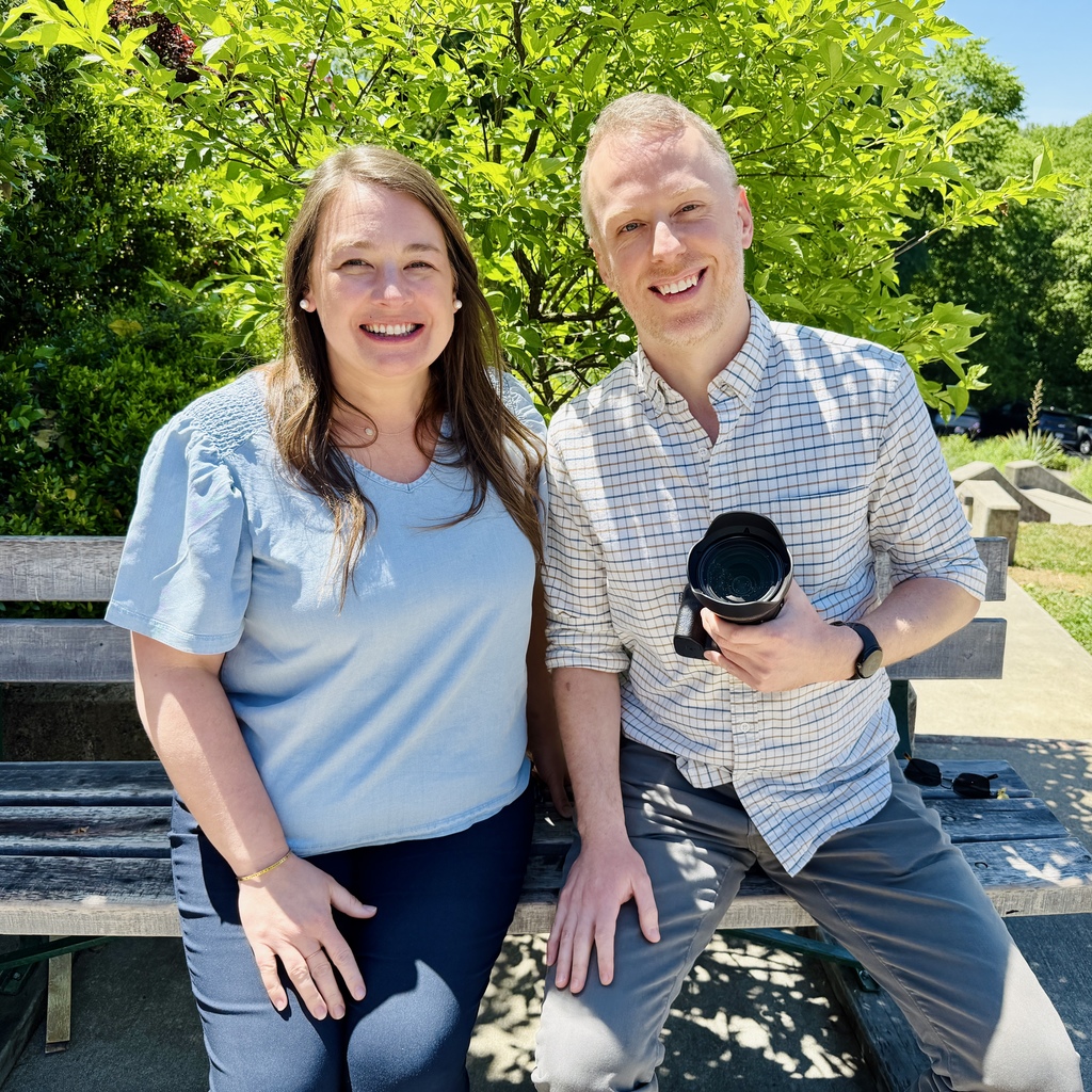Emily and James sitting on a bench with green bush behind them.  James is holding a camera.