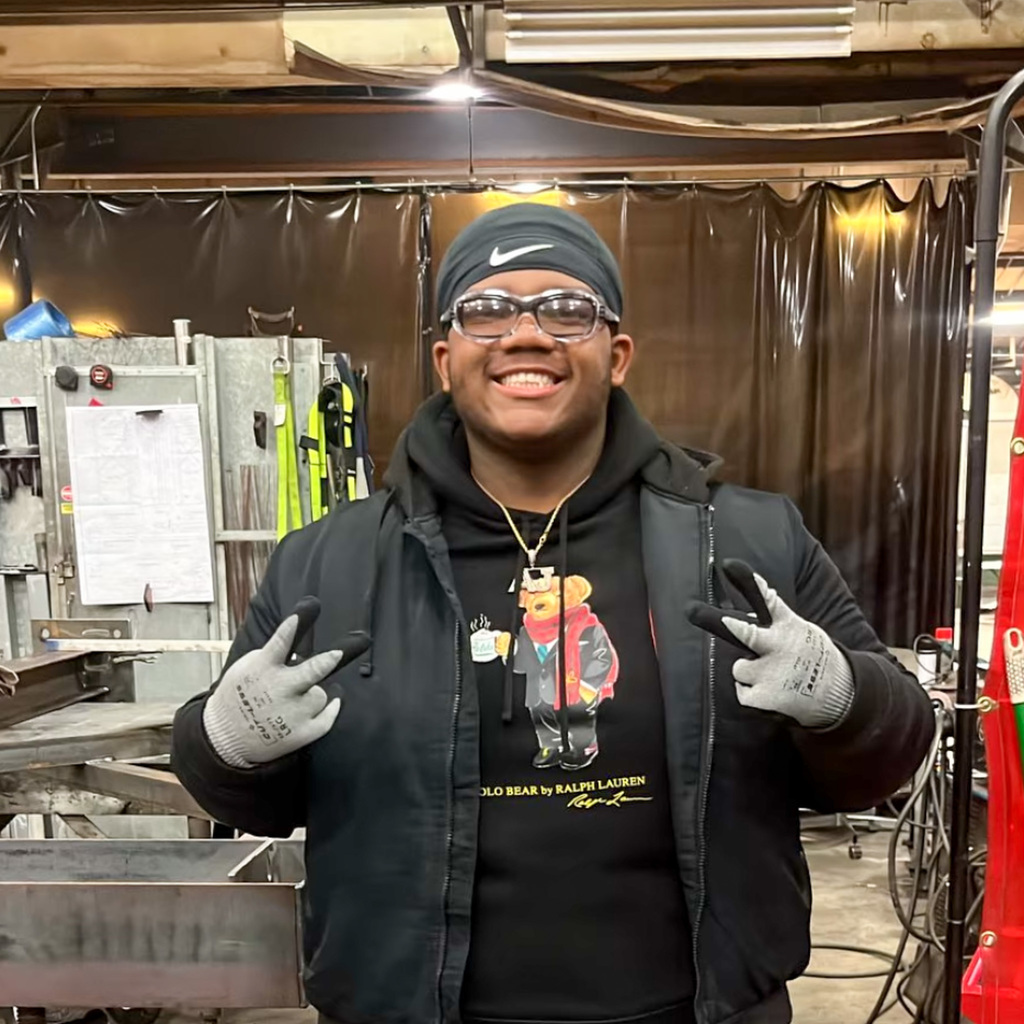 A high school senior wearing safety glasses and a black headband gives double peace signs in an industrial work space