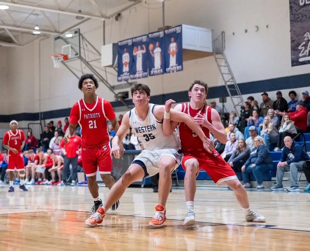 Three high school basektball athletes, two wearing red Patriots jerseys and one wearing a white Western jersey, jostle for a rebound