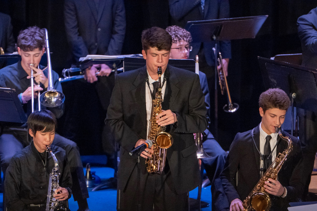 A high school jazz band plays on stage under blue lights. They are all wearing black formal attire. The front three are playing saxophones and a few others in the back have trumpets.