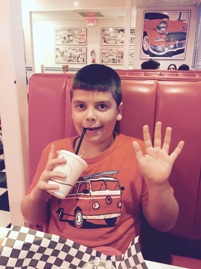 A young boy wearing an orange RV t-shirt smiles and waves while seated in a diner restaurant