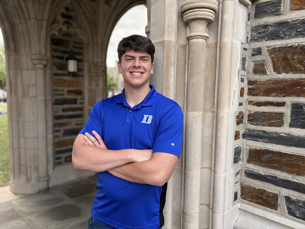 A high school senior wearing a Duke blue polo shirt leans against a stone archway in a brick outdoor hallway