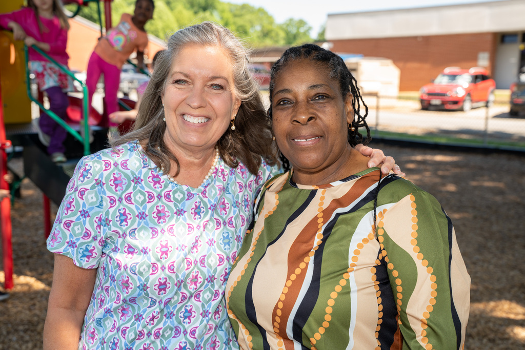 A warm portrait of two women smiling together on a playground. The woman on the left has grey hair and a floral blue blouse, and the woman on the right has dark braided hair and a green and brown patterned shirt.