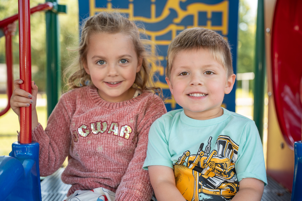A joyful portrait of two children sitting together on a playground structure. The girl on the left wears a pink "Hogwarts" sweater, and the boy on the right wears a light blue t-shirt with a construction vehicle graphic.