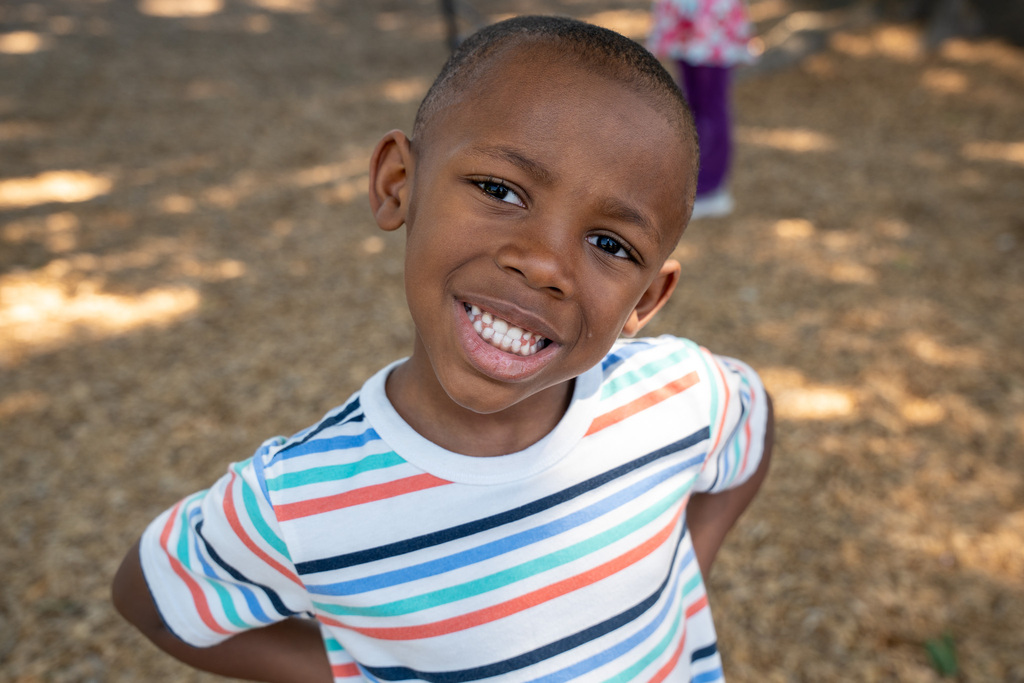 A close-up, high-angle portrait of a young boy with a bright smile, wearing a colorful striped t-shirt. He is standing outside on a woodchip-covered playground.