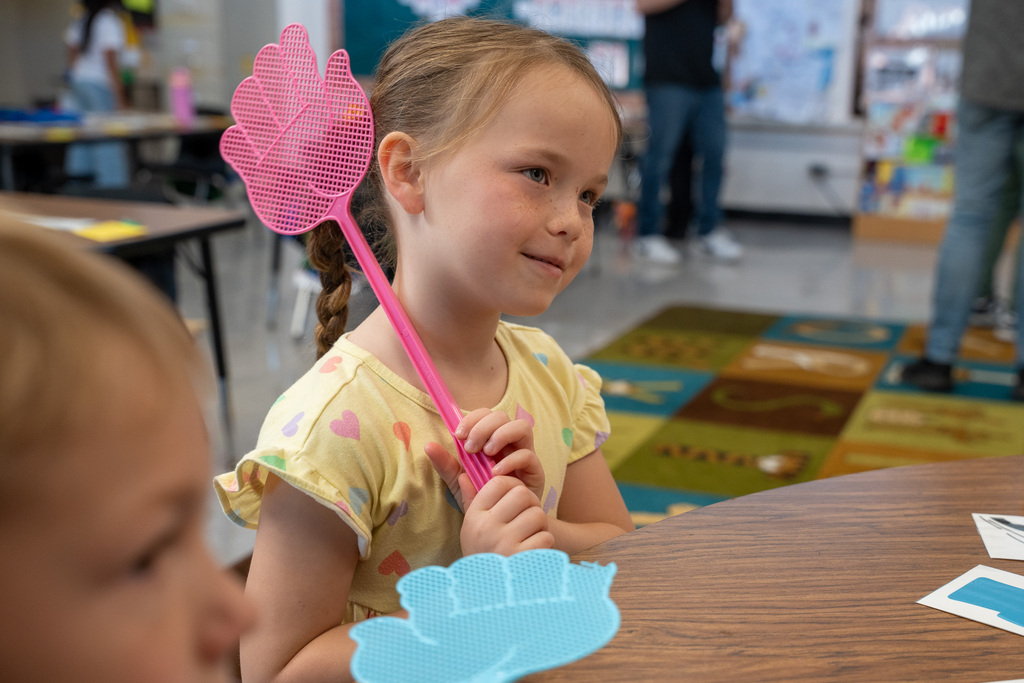 A close-up of a young girl with a braid, smiling and looking off-camera while holding a pink, hand-shaped fly swatter. A blue fly swatter rests on the table in the foreground.