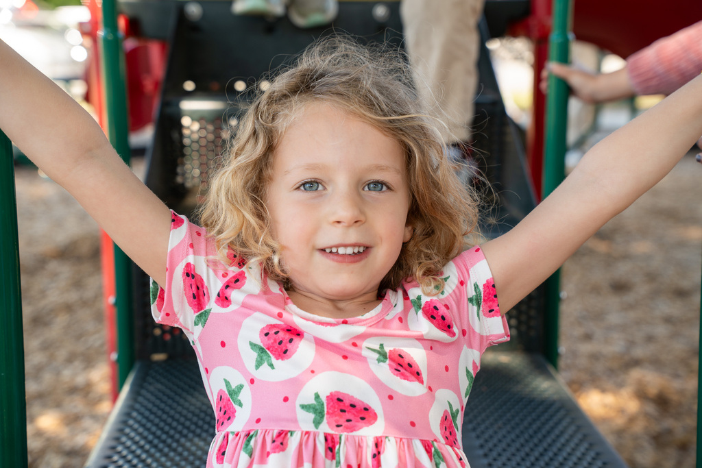 A young girl with curly blonde hair and blue eyes smiles at the camera while sitting on a playground slide. She is wearing a pink dress patterned with watermelons and has her arms raised out to the sides.