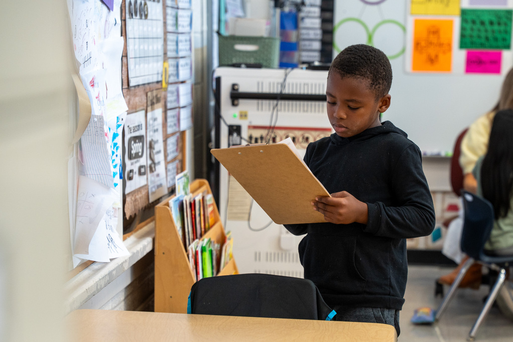 A young boy in a black hoodie stands in a classroom, looking down and concentrated as he holds a wooden clipboard. Behind him are bookshelves and various student projects pinned to the wall.