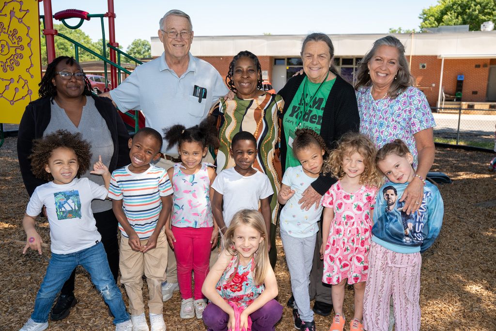 A group photo on a sunny playground featuring five adults and nine young children. Everyone is smiling and posed closely together in front of a colorful climbing structure and a brick school building.