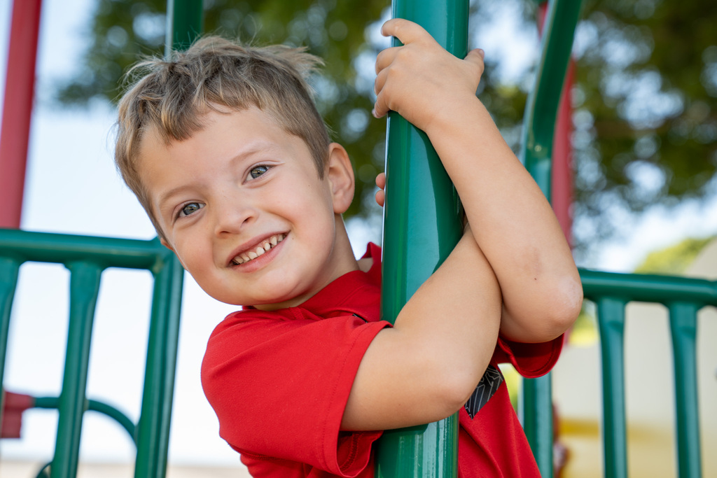 A cheerful young boy in a red shirt leans against a green metal playground pole, smiling broadly at the camera with his arms wrapped around the post.