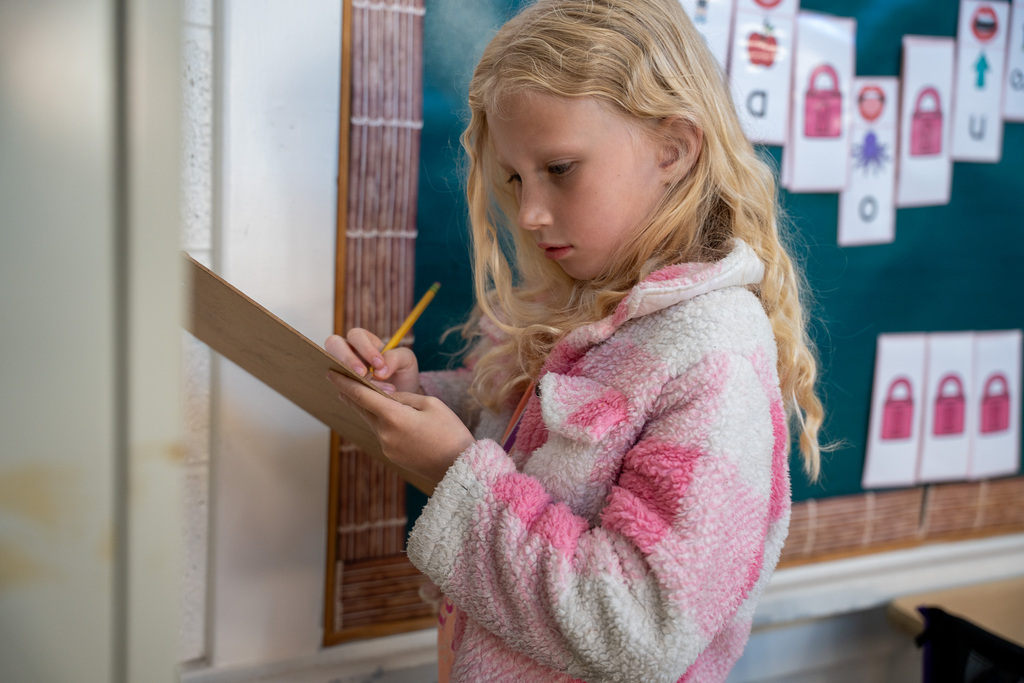 A young girl with long blonde hair and a pink patterned fleece jacket focuses intently on writing on a clipboard. In the background, a green bulletin board displays literacy lessons with images of locks and apples.