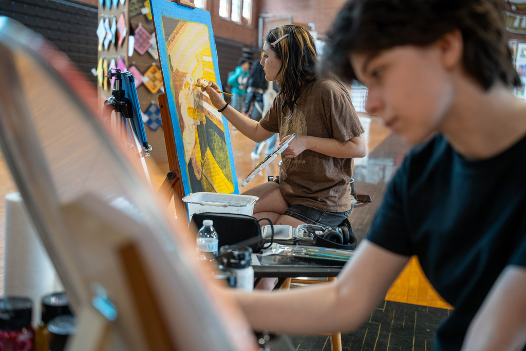 A student in a brown t-shirt paints a colorful landscape on an easel, while another student is seen in soft focus in the foreground.