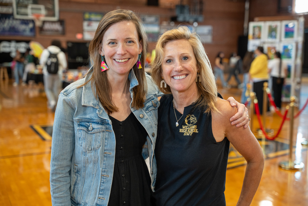 Two women stand together smiling in a gymnasium. One wears a denim jacket and colorful earrings; the other wears a black "Williams High School Staff" shirt.