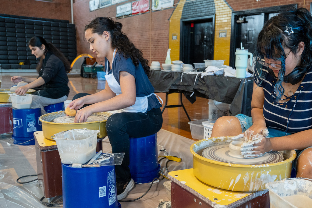 Three students sit at pottery wheels in a gymnasium, focused on shaping clay. The student in the foreground uses both hands to mold a bowl.