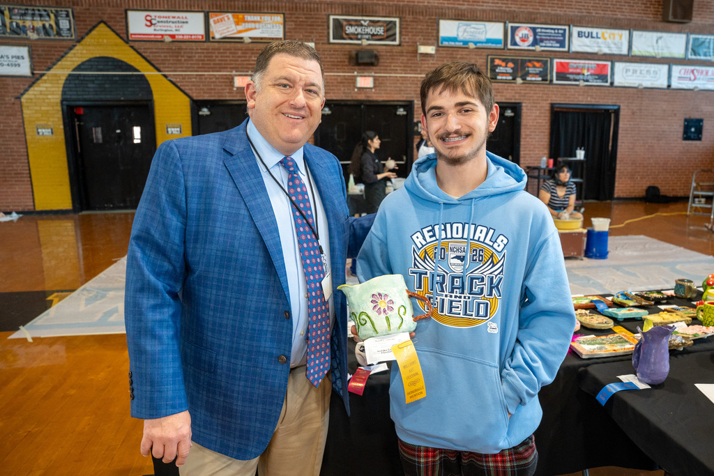 A smiling student in a blue "Regionals 2026 Track and Field" hoodie holds a decorated ceramic pitcher next to a man in a blue suit jacket.