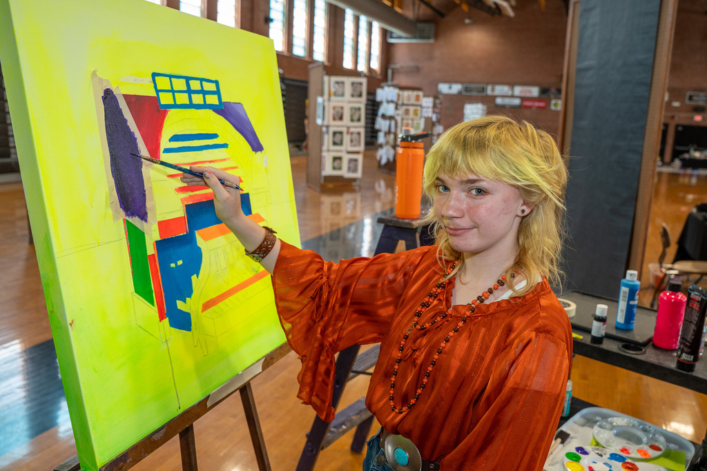 A young artist with blonde hair and an orange blouse focuses on a large, vibrant yellow canvas, applying purple paint with a brush.