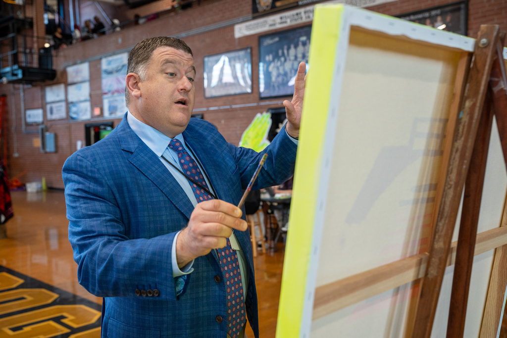 A man in a blue patterned suit and tie gestures with his hands while looking at the back of a large painting canvas on an easel.