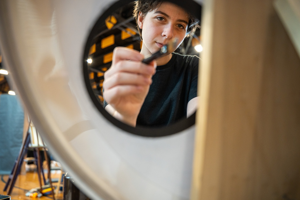 A unique, circular perspective through a prop showing a student artist holding a paintbrush toward the camera.