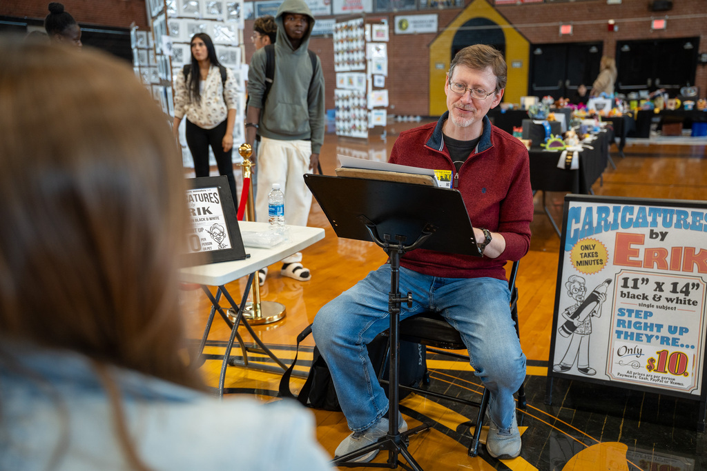 An artist named Erik sits at a drawing stand in a gym, preparing to draw a caricature for a student. A sign next to him advertises "$10 Caricatures."