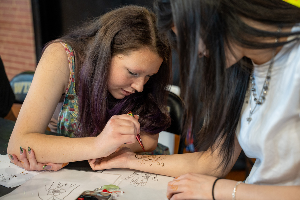 A student carefully applies a henna tattoo to the forearm of another student seated closeby