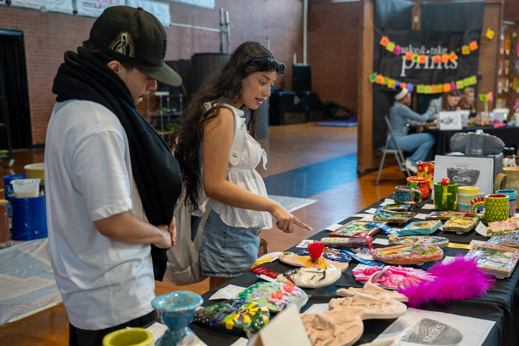 Two students stand at a long table covered in black cloth, admiring a diverse collection of colorful ceramic masks and pottery on display.