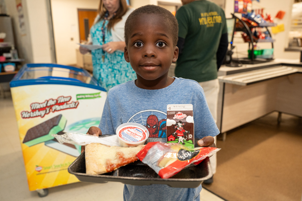 A young boy looks directly at the camera with wide, excited eyes while balancing a lunch tray that holds a slice of pizza, a bag of apple slices, chocolate milk, and a cup of celebratory ice cream.