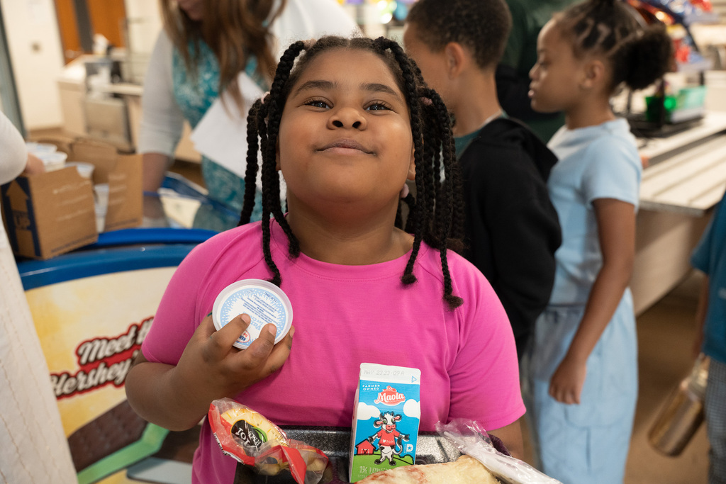 A young girl with braided hair looks up and smiles contentedly while holding a small cup of ice cream and a school lunch tray featuring pizza and milk.