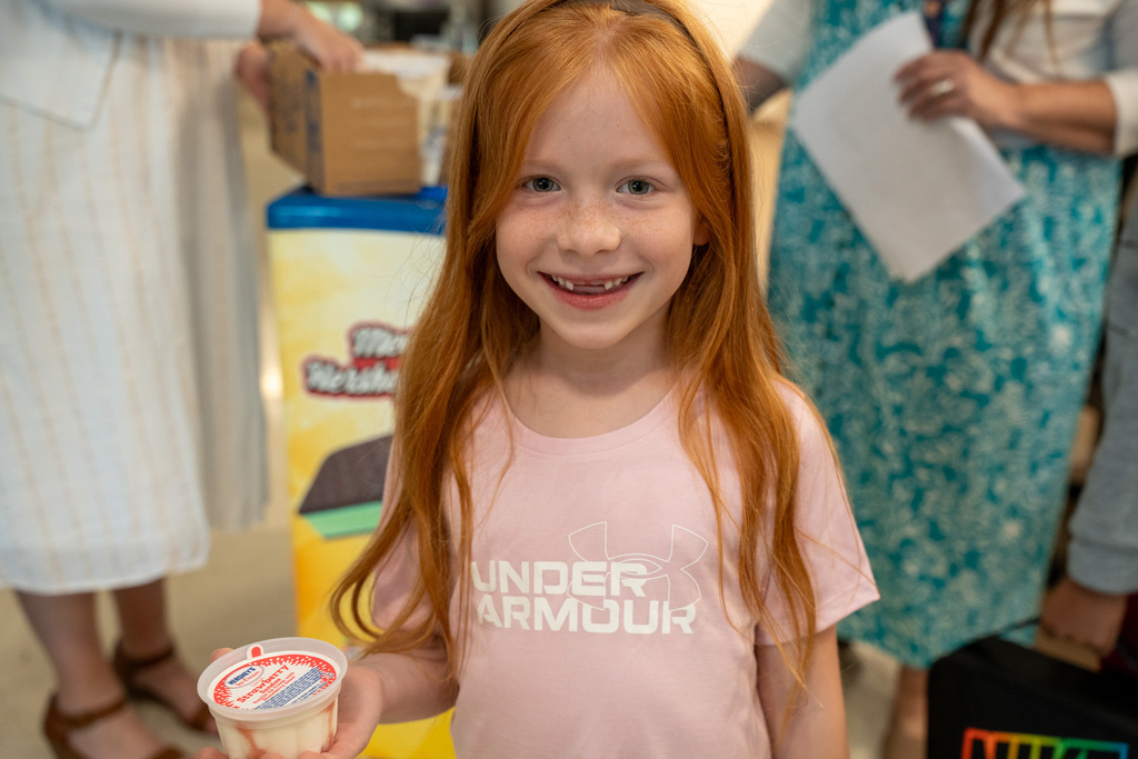 A smiling young girl with long red hair holds up a small cup of strawberry swirl ice cream, celebrating the school's attendance achievement.
