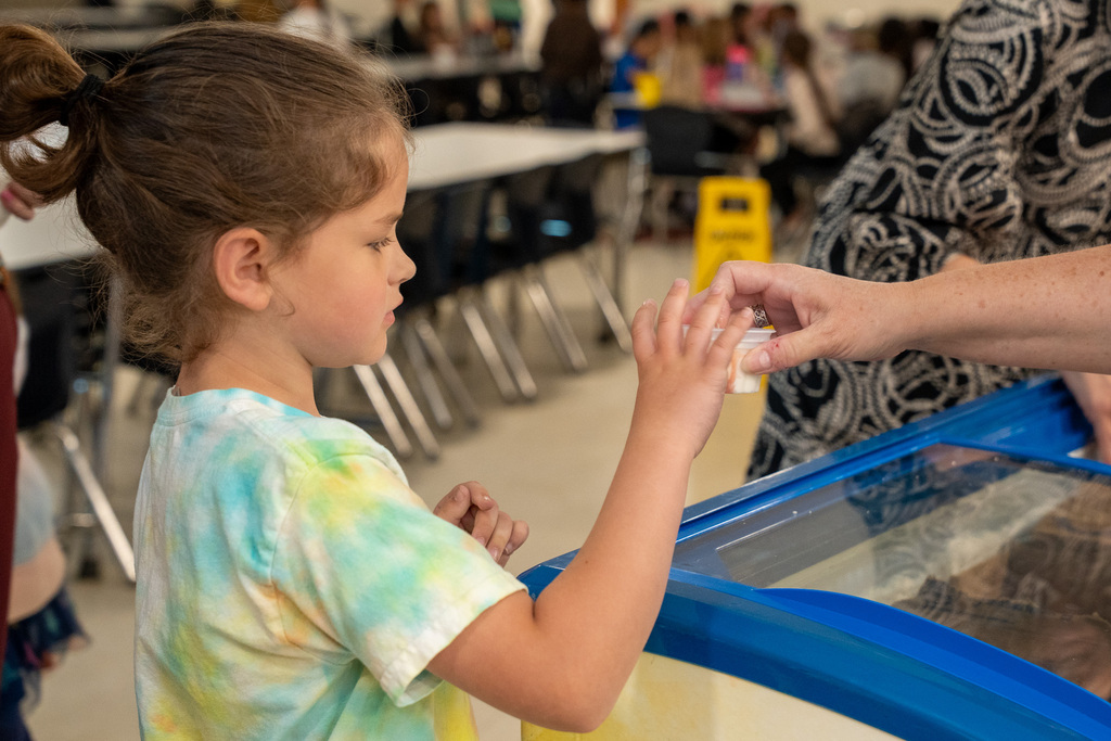 A close-up side profile of a young student with her hair in a bun reaching out to receive a surprise ice cream treat from a staff member’s hand over an ice cream freezer.