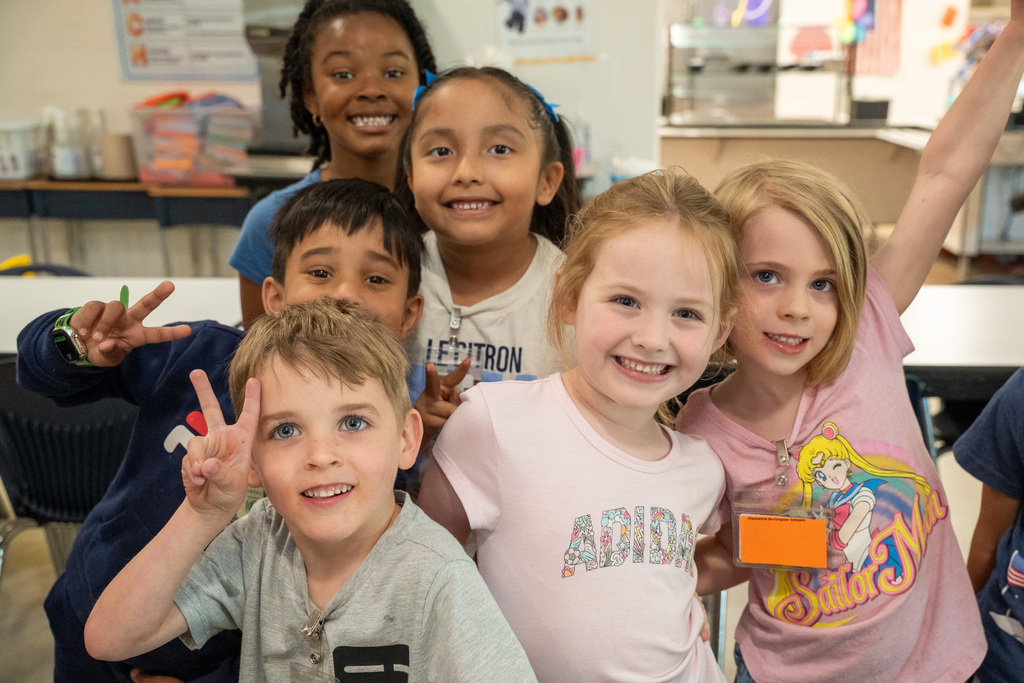 A joyful group of six young students huddle together for a close-up photo, smiling broadly and making peace signs for the camera in the school cafeteria.