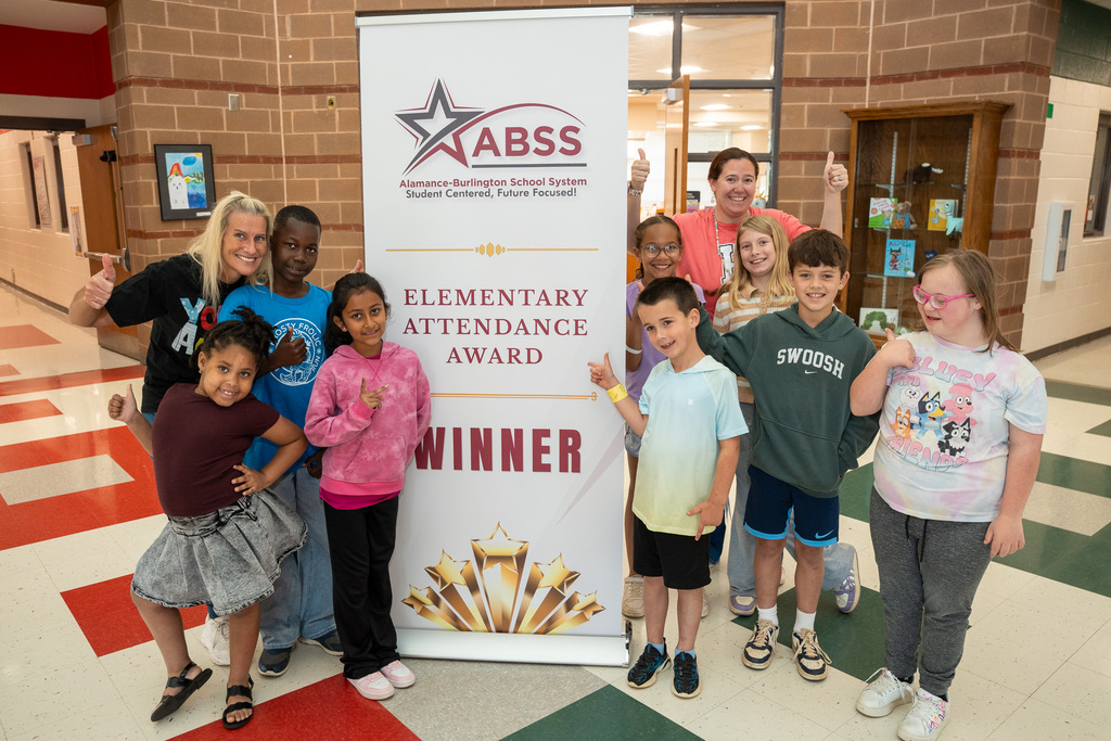 A group of smiling elementary students and two staff members stand proudly on either side of a large white banner that reads "ABSS Elementary Attendance Award Winner." Everyone is giving a thumbs-up or peace sign in a bright school hallway.