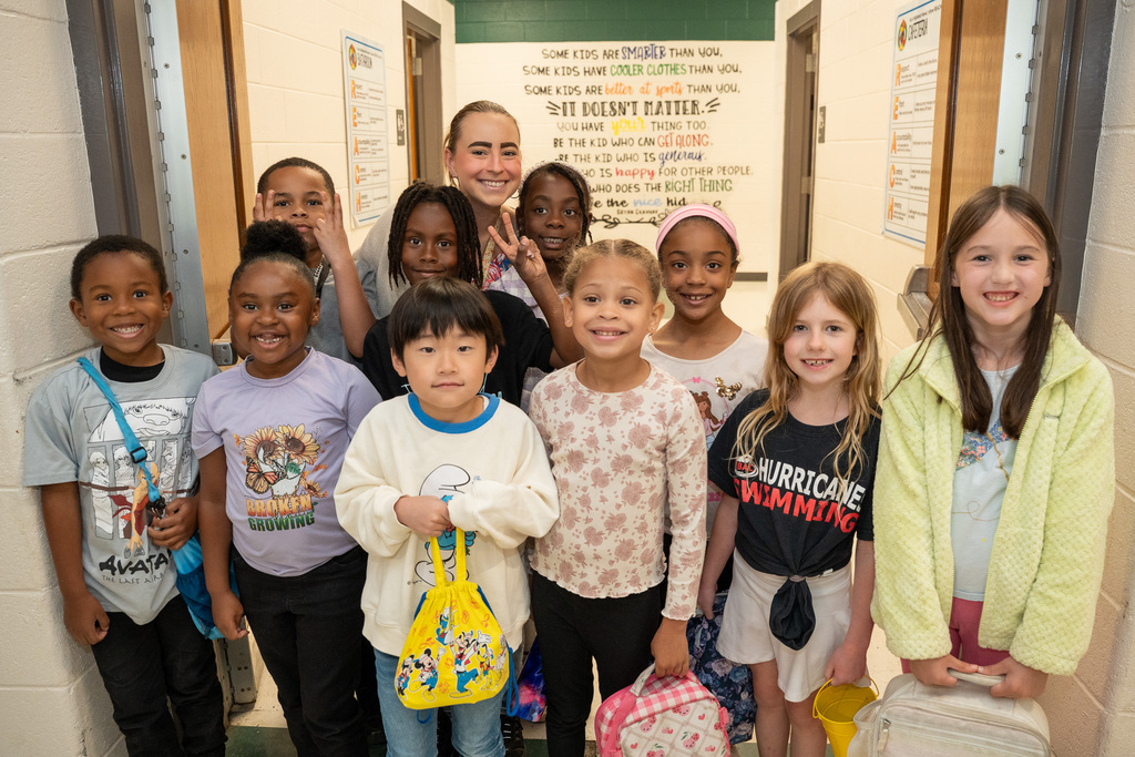 A diverse group of ten elementary students and one teacher pose together in a school hallway. Behind them, a wall mural features an inspirational quote about kindness and character.