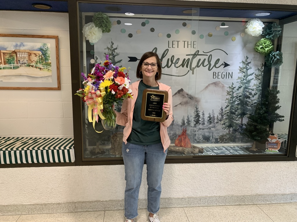 Nurse Donna Apple holding flowers and a plaque in front of a Let the Adventure Begin school display