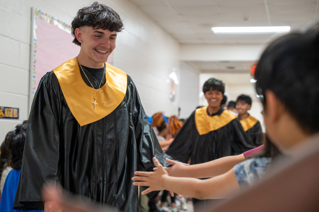 A male graduate in a black gown with a gold stole walks down a school hallway, smiling as he looks down to high-five elementary school students who are reaching their hands out from the side.