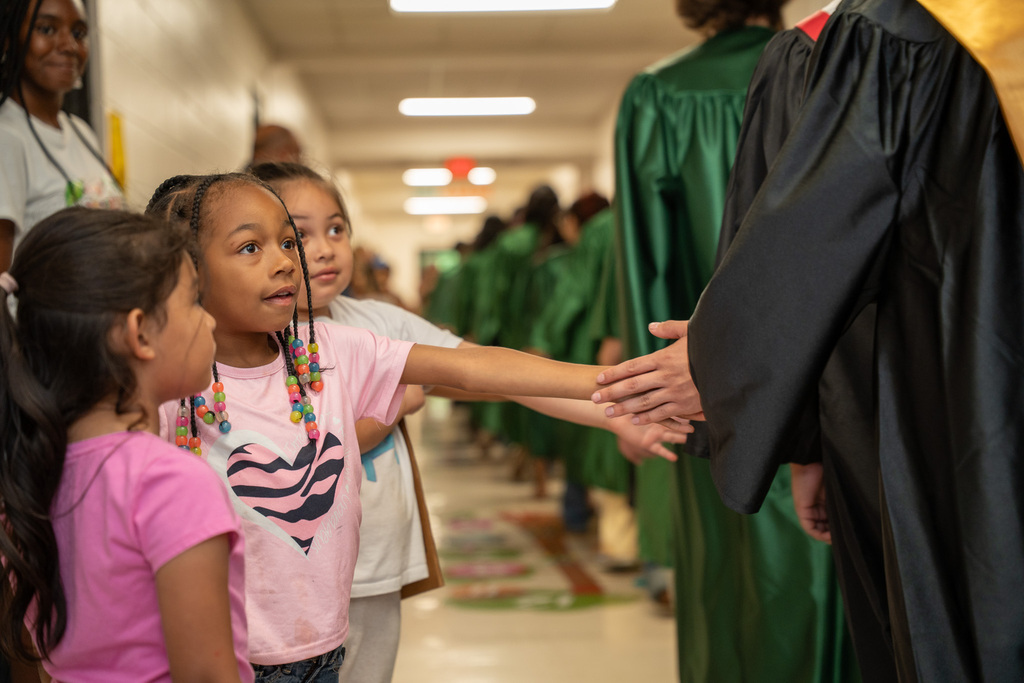 A young elementary school girl in a pink shirt with a heart pattern reaches her hand out eagerly to high-five passing high school graduates lined up in a school hallway during a graduation walk.