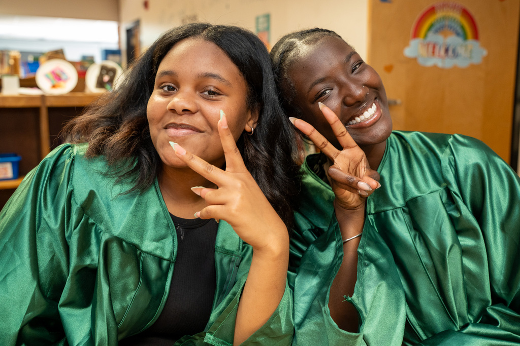 Close-up portrait of two young female graduates wearing shiny green gowns sitting inside a classroom. Both are smiling brightly at the camera while making peace signs with their hands near their faces.