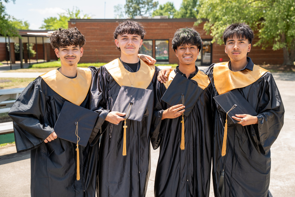 Four smiling male graduates stand shoulder-to-shoulder outdoors in front of a brick school building. They are wearing black graduation gowns with gold stoles and each holds a black mortarboard cap with a gold tassel in front of them. The two graduates in the center have their arms around each other's shoulders.