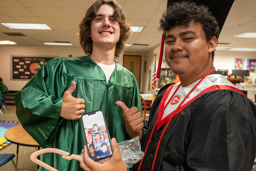 Two male graduates smile for a photo inside a classroom. The graduate on the left is in a green gown and gives a thumbs-up. The graduate on the right, wearing a black gown with a red and white stole, holds up a smartphone showing an old childhood photo of them as young kids in elementary school.
