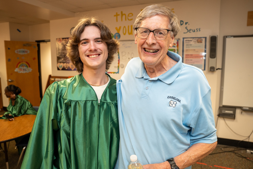 Inside a classroom, a young male graduate with wavy brown hair wearing a green graduation gown smiles warmly with his arm around an older man with glasses. The older man is smiling broadly and wearing a light blue polo shirt with a University of North Carolina "UNC" logo.
