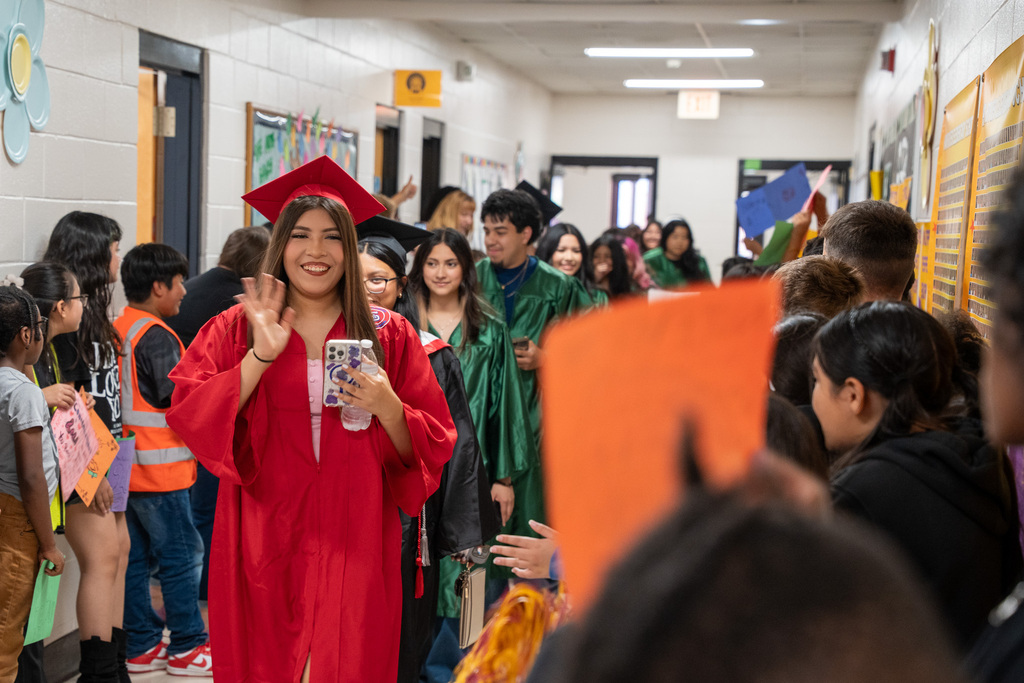 A female graduate in a red cap and gown waves happily to the camera as she leads a procession of graduates down a crowded elementary school hallway. Younger students line the walls, holding up colorful signs to celebrate them.