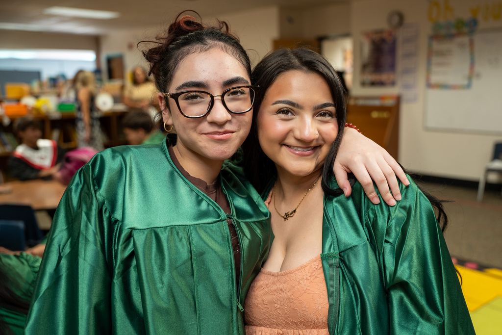 Close-up of two smiling young women wearing green graduation gowns posing together indoors. The woman on the right has her arm draped affectionately over the shoulder of the woman on the left, who is wearing glasses.