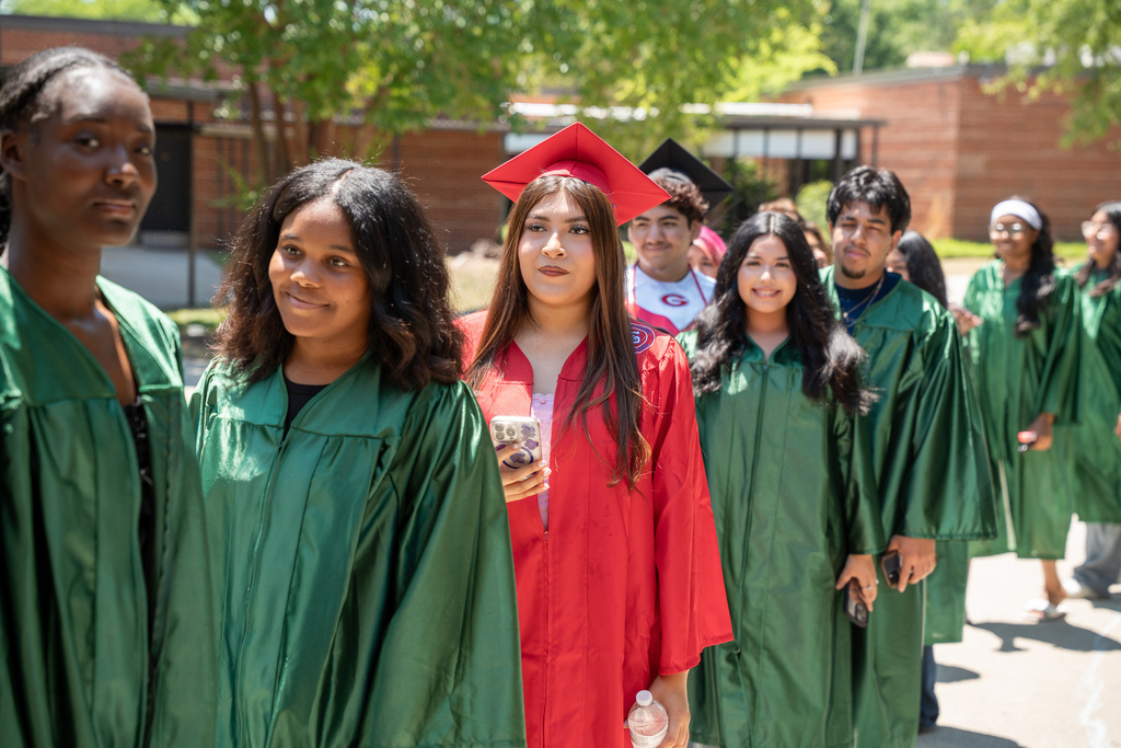 A line of high school graduates in green, red, and black gowns walk in a procession outdoors on a sunny day. A female graduate in a red gown and cap walks near the front of the line looking ahead, while others follow behind her.