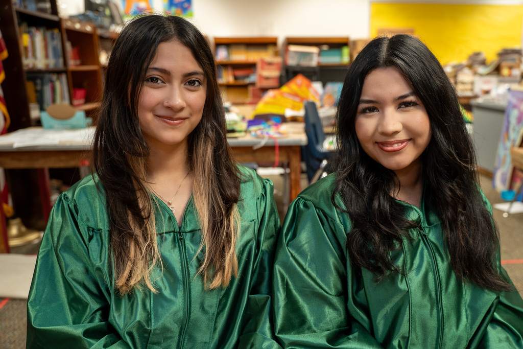 Two young women wearing green graduation gowns sit side-by-side inside a classroom, smiling pleasantly directly at the camera.