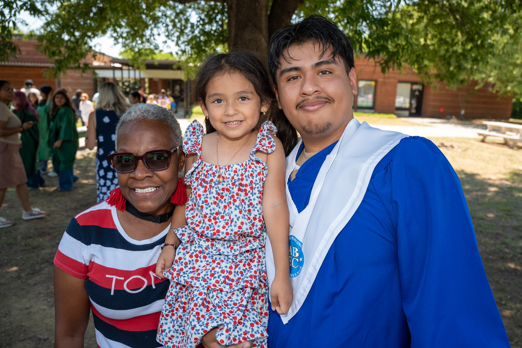 An outdoor group photo of a male graduate in a blue cap and gown holding a young girl in a patterned dress, alongside an older woman wearing sunglasses, red earrings, and a striped shirt. They are all smiling outside a brick school building with other graduates blurred in the background.