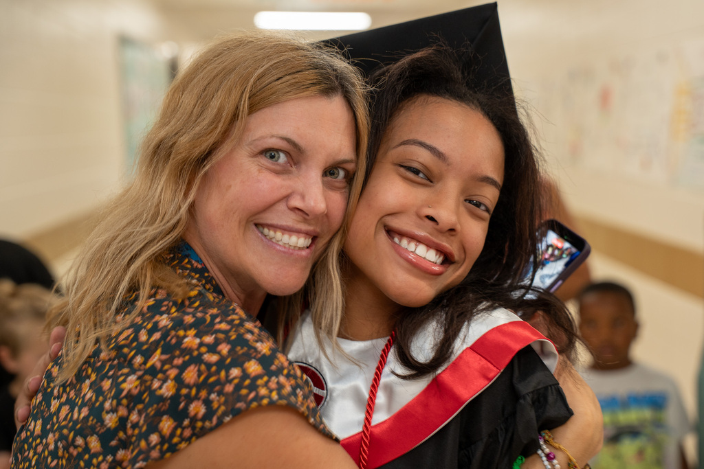 Close-up portrait of a smiling adult woman hugging a teenage girl wearing a black graduation gown, red stole, and a black mortarboard cap. Both are smiling warmly at the camera inside a school hallway.