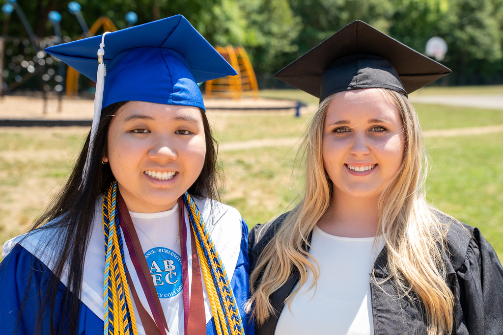 A medium portrait of two female graduates standing side-by-side outdoors. The graduate on the left wears a blue cap and gown with an "ABEC" logo stole and multiple honor cords. The graduate on the right wears a black cap and gown over a white dress. Both are smiling at the camera.