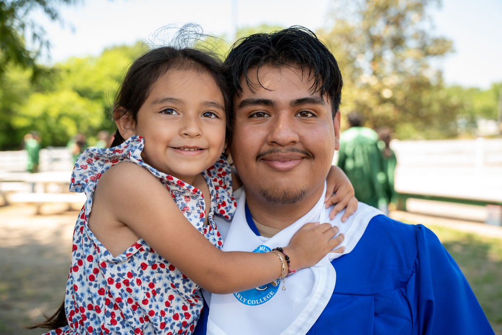 Close-up outdoor portrait of a male graduate in a blue gown and white stole with an "Early College" logo. A young girl in a patterned red and blue dress is wrapped around his neck, hugging him tightly as both smile at the camera.
