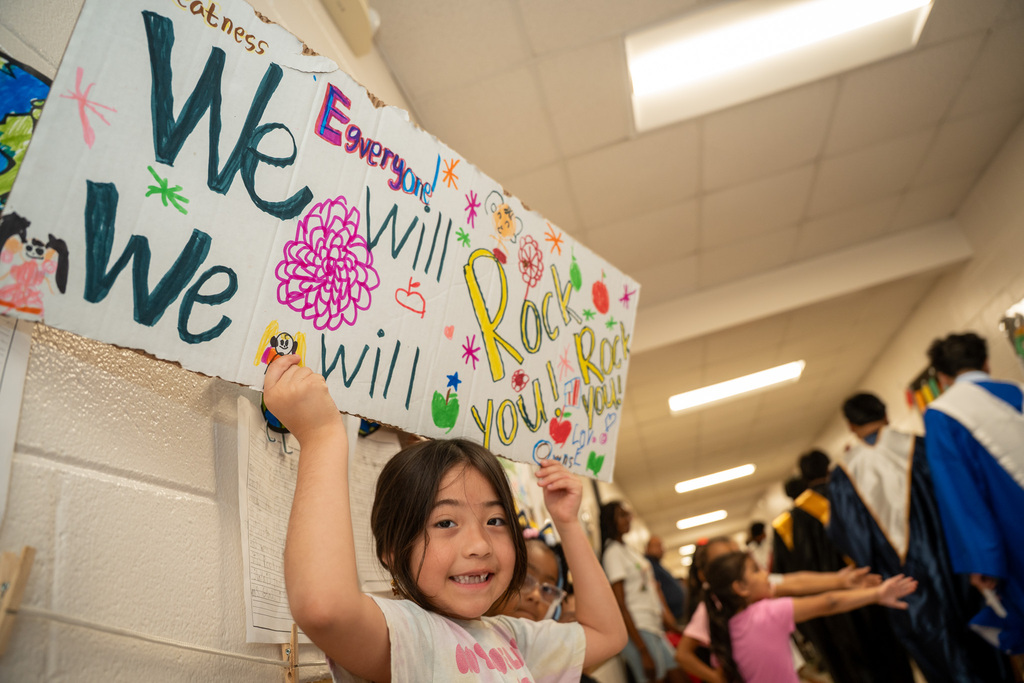 A young elementary school girl with a wide smile stands in a school hallway, holding up a large cardboard sign over her head. The colorful, hand-drawn sign reads "We Will Rock You!" and "Everyone!" surrounded by drawings of flowers and apples. High school seniors in graduation gowns walk down the hallway behind her as other young children reach out for high-fives.
