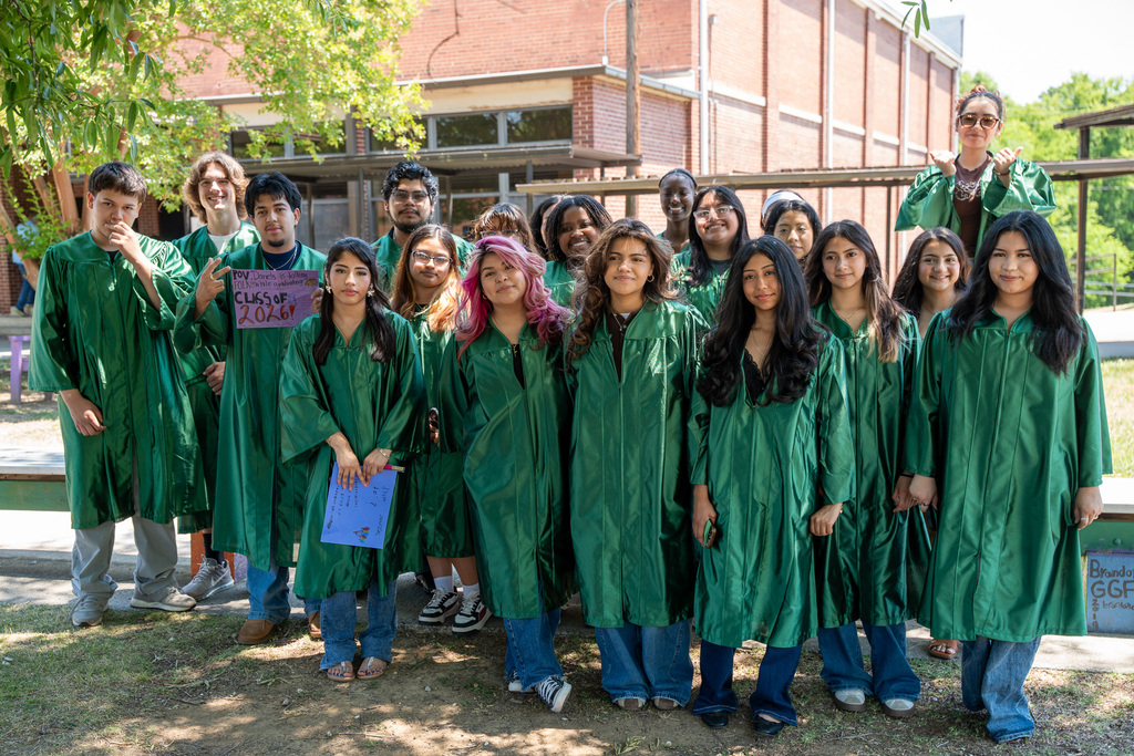 A group photo of about fifteen high school seniors posing outdoors in front of a brick school building. They are all wearing shiny green graduation gowns. One student holds a small pink sign that reads "Class of 2026."