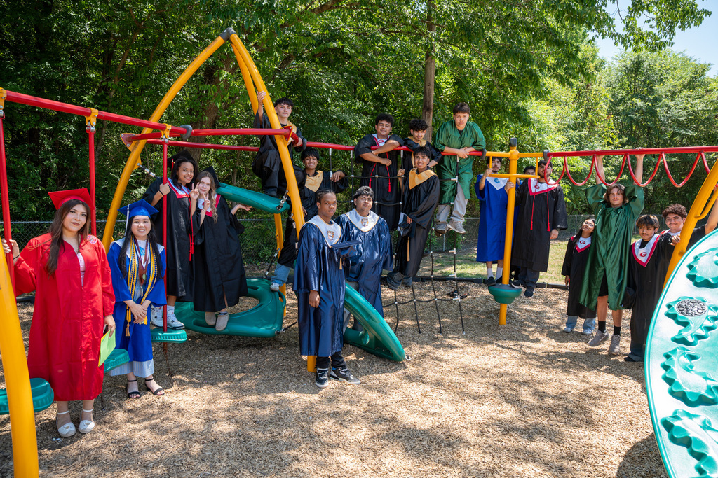 A wide outdoor shot of a diverse group of high school graduates in various colored gowns—including red, blue, green, and black—posing playfully on and around a colorful metal playground structure. Trees line the background under a bright, sunny sky.